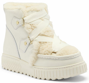 A cream-colored, ankle-length boot with a thick, ridged platform sole, featuring a white ribbon-style lace-up closure over a shearling-lined front and gold-tone metal hardware.