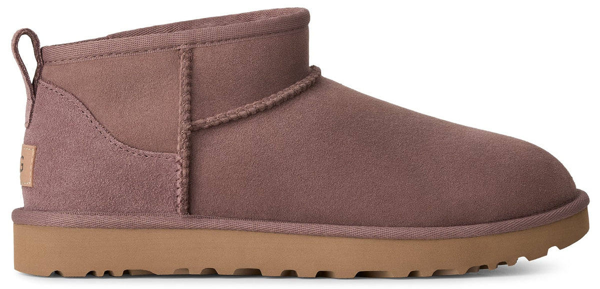 A mauve-colored suede ankle boot with a tan, thick, treaded rubber sole, showing a pull tab on the back and a small rectangular patch with the text UGG on the heel.