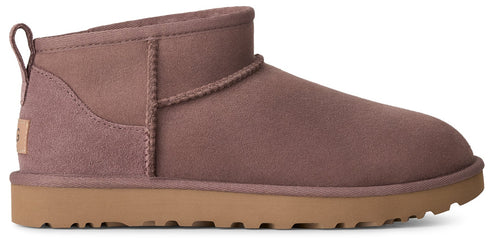 A mauve-colored suede ankle boot with a tan, thick, treaded rubber sole, showing a pull tab on the back and a small rectangular patch with the text UGG on the heel.