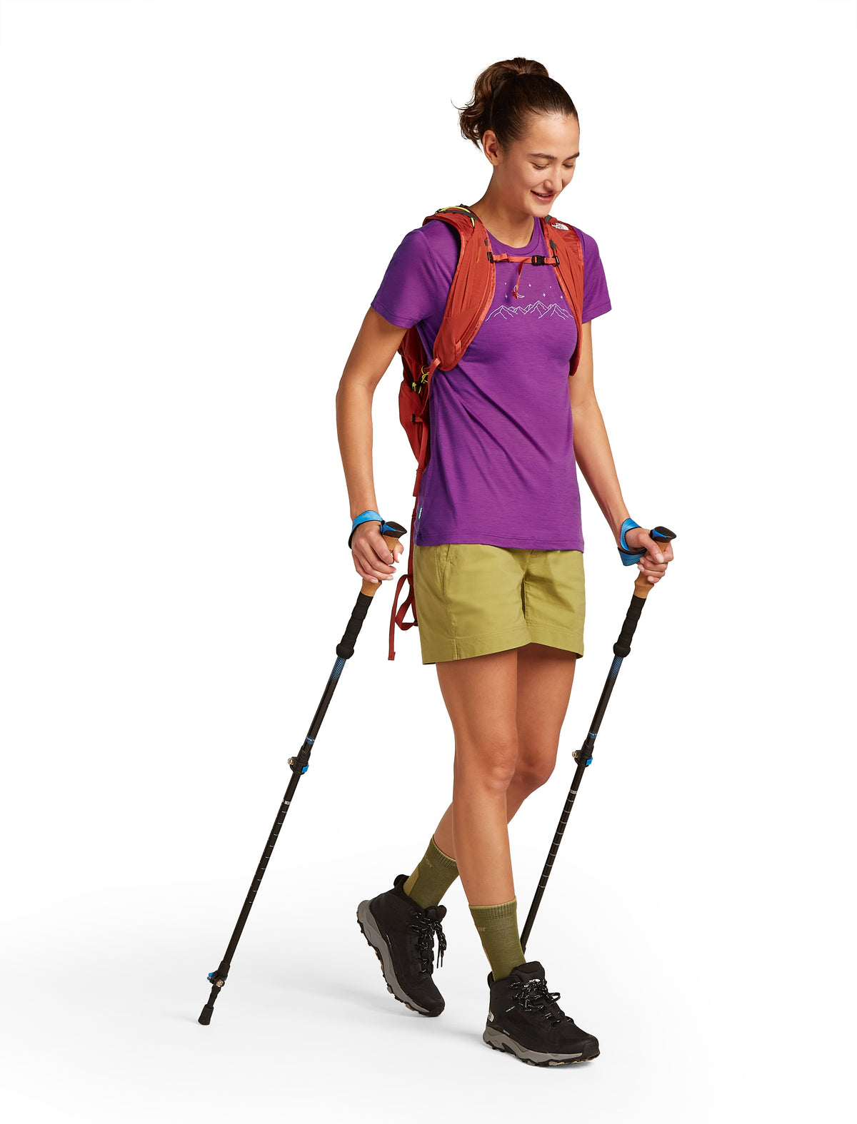 A woman with trekking poles wearing an orange pack, purple tee shirt, green shorts, olive socks, and black hiking boots.
