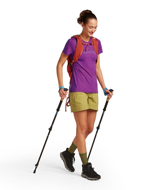 A woman with trekking poles wearing an orange pack, purple tee shirt, green shorts, olive socks, and black hiking boots.