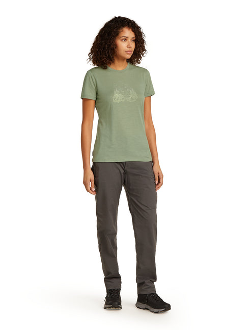 A woman wearing a green t-shirt with a graphic design and charcoal-colored pants and black shoes stands facing right.