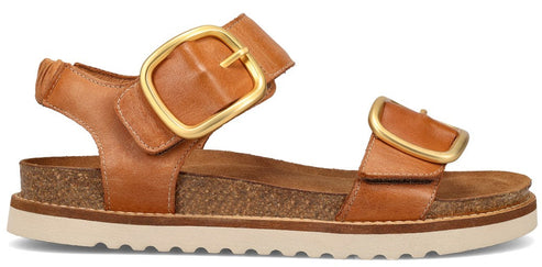 A tan sandal with two straps, each with a large gold buckle.
