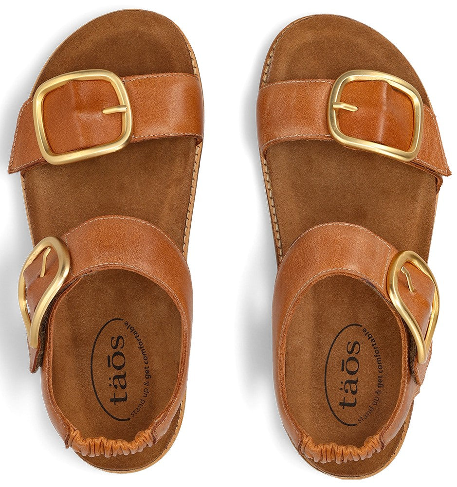 Two leather sandals with large gold buckles and “tāos” text on the soles.