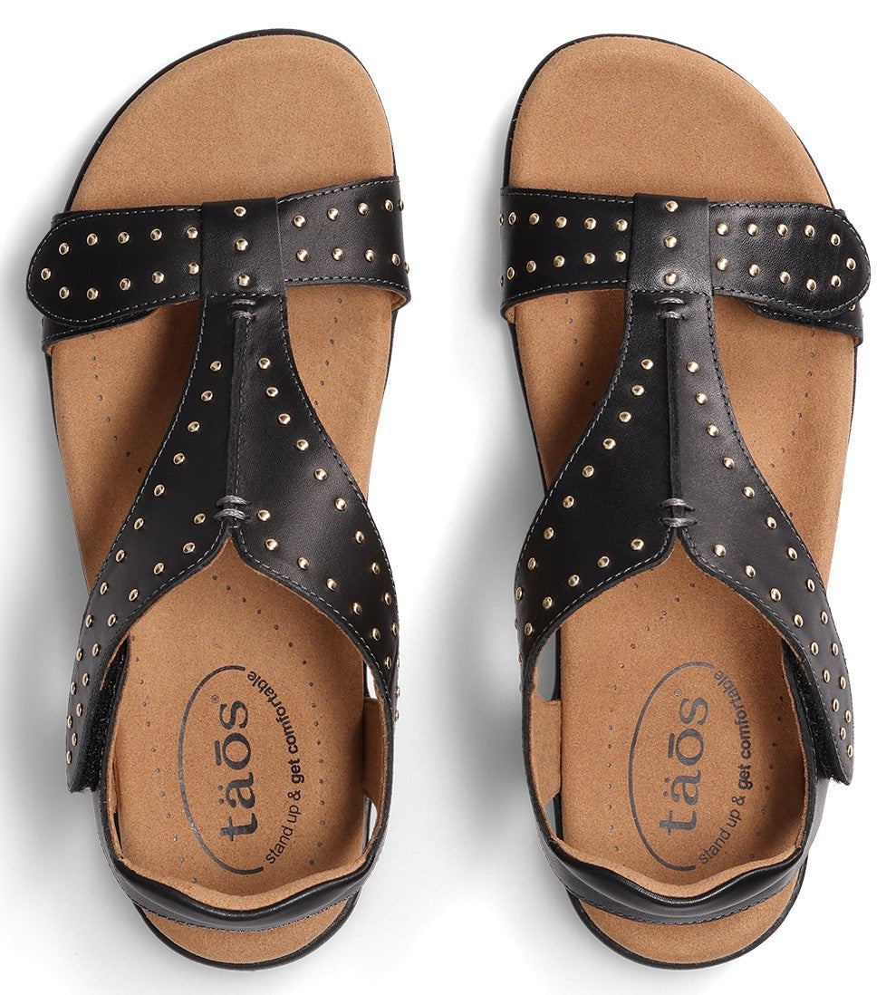 Pair of black T-strap sandals with decorative silver studs and "taos" written on the insole.