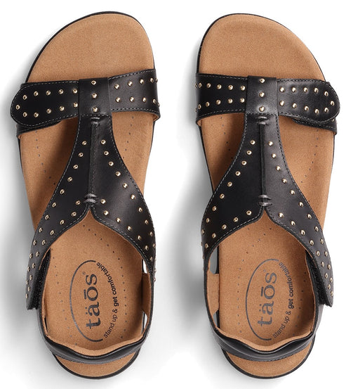 Pair of black T-strap sandals with decorative silver studs and "taos" written on the insole.