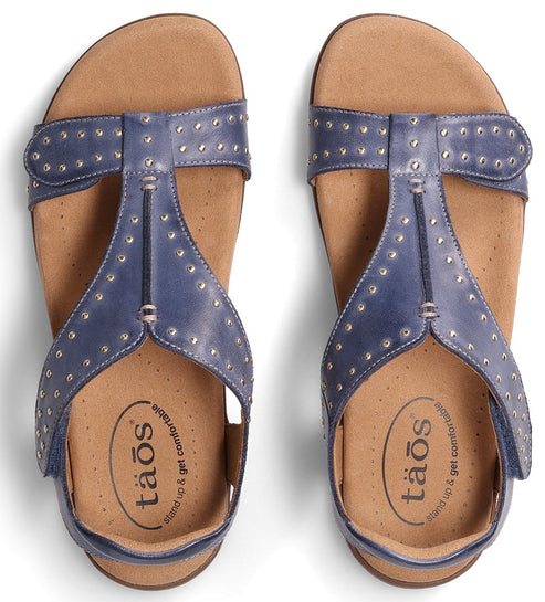 A pair of blue T-strap sandals with gold rivets and a cushioned insole that reads "tãos stand up & get comfortable".