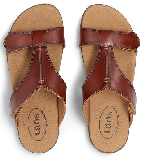 Pair of brown sandals with the text "taos stand up & get comfortable" printed on the inner sole.