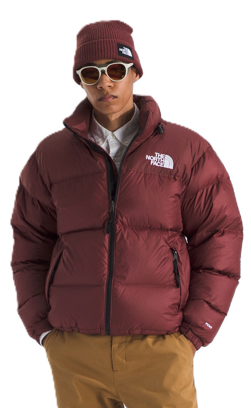 A person wearing a maroon puffer jacket and matching beanie, with light-colored pants and sunglasses. A logo is on the jacket's chest.