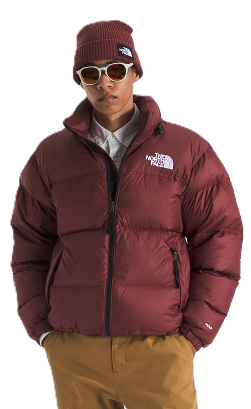 A person wearing a maroon puffer jacket and matching beanie, with light-colored pants and sunglasses. A logo is on the jacket's chest.