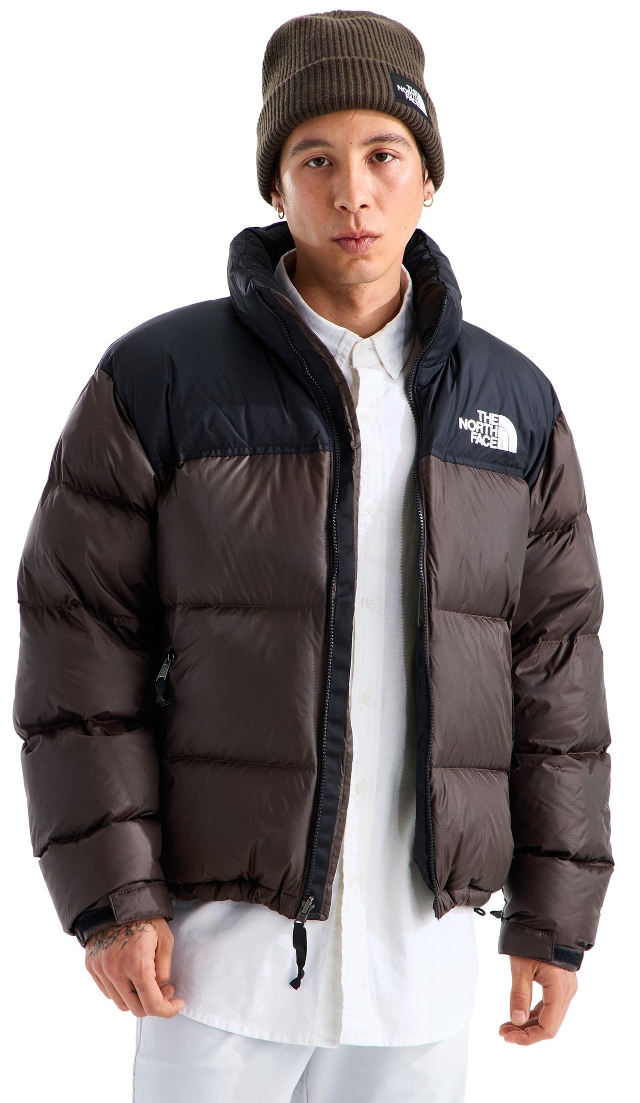 A man wearing a brown and black puffer jacket with a high collar, a white button-up shirt underneath, and a brown knit beanie with a logo patch.