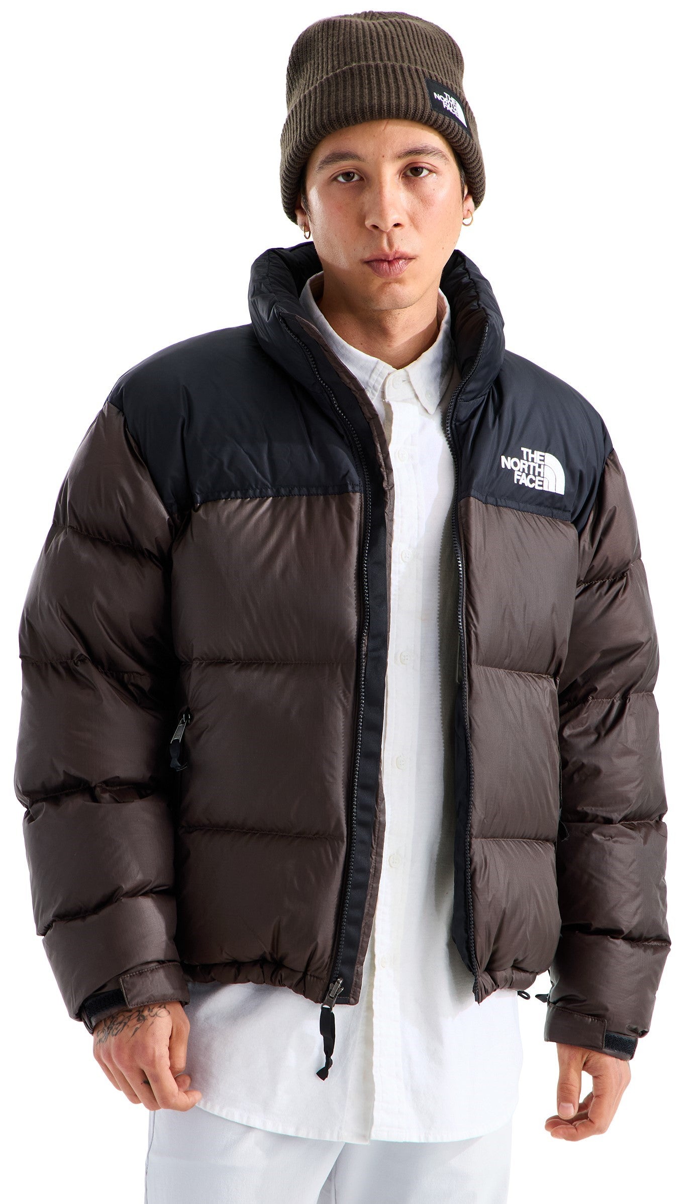 A man wearing a brown and black puffer jacket with a high collar, a white button-up shirt underneath, and a brown knit beanie with a logo patch.