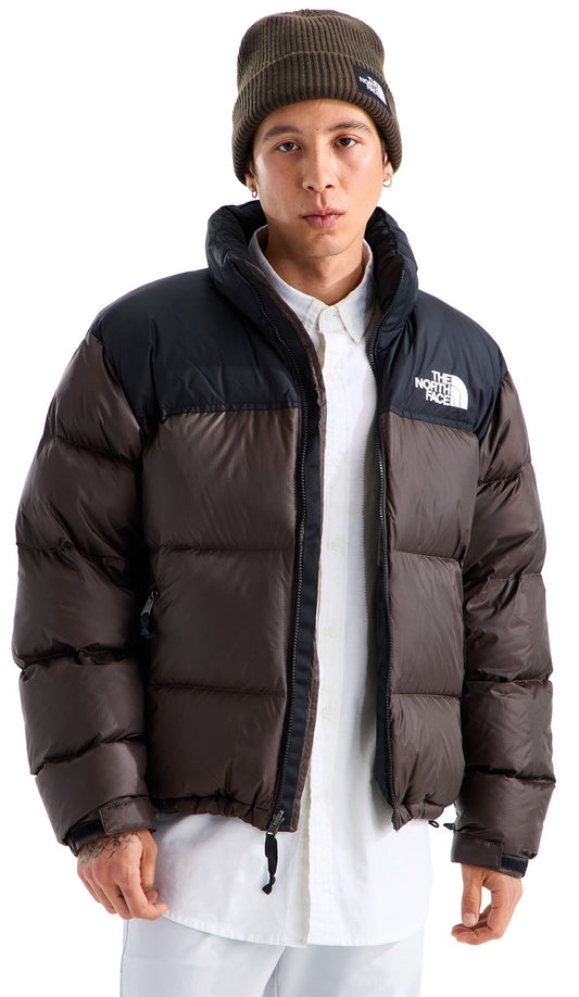 A man wearing a brown and black puffer jacket with a high collar, a white button-up shirt underneath, and a brown knit beanie with a logo patch.