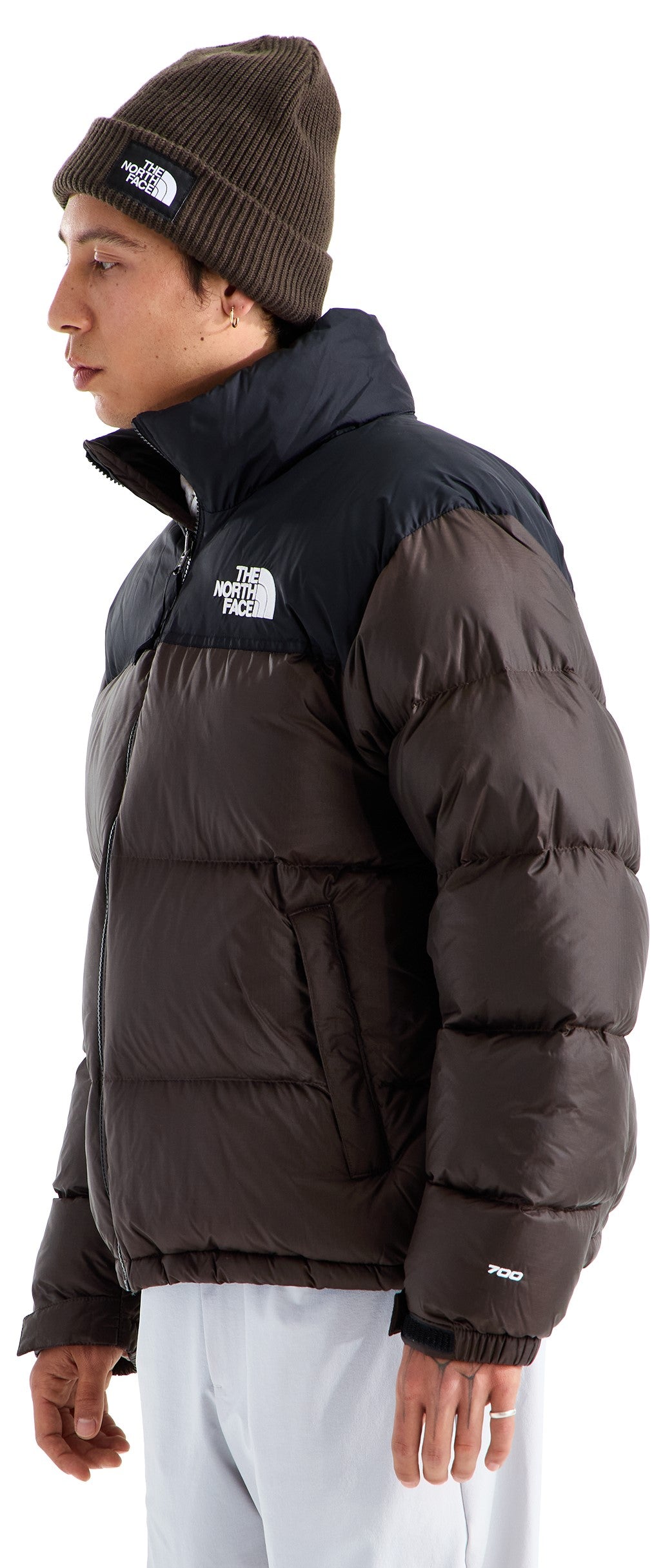 A person in profile wearing a dark brown puffer jacket with a black yoke, collar, and The North Face logo on the chest, along with a matching dark brown beanie and light-colored pants.