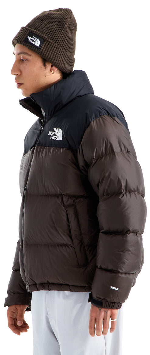 A person in profile wearing a dark brown puffer jacket with a black yoke, collar, and The North Face logo on the chest, along with a matching dark brown beanie and light-colored pants.