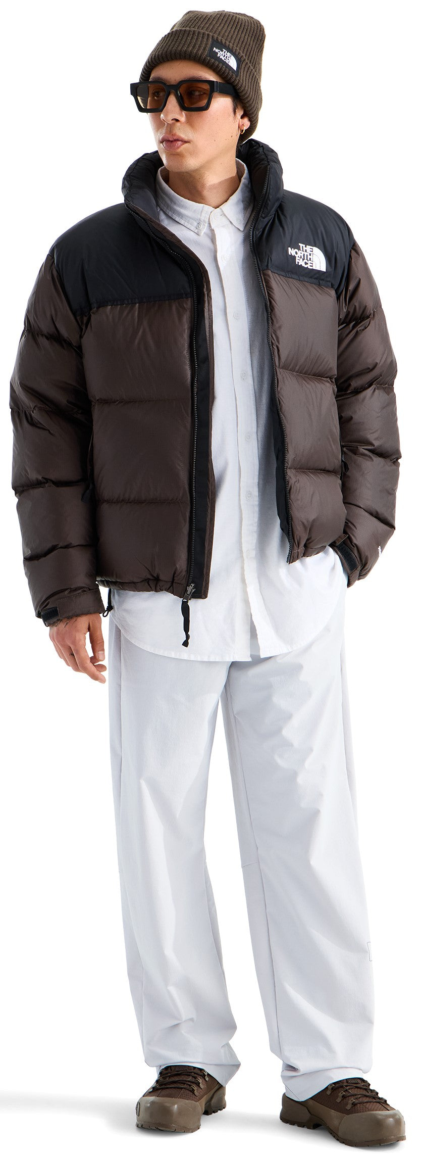 A male model wearing a dark brown and black puffer jacket, a white button-down shirt, loose-fitting light-colored pants, a brown beanie, and dark sunglasses.