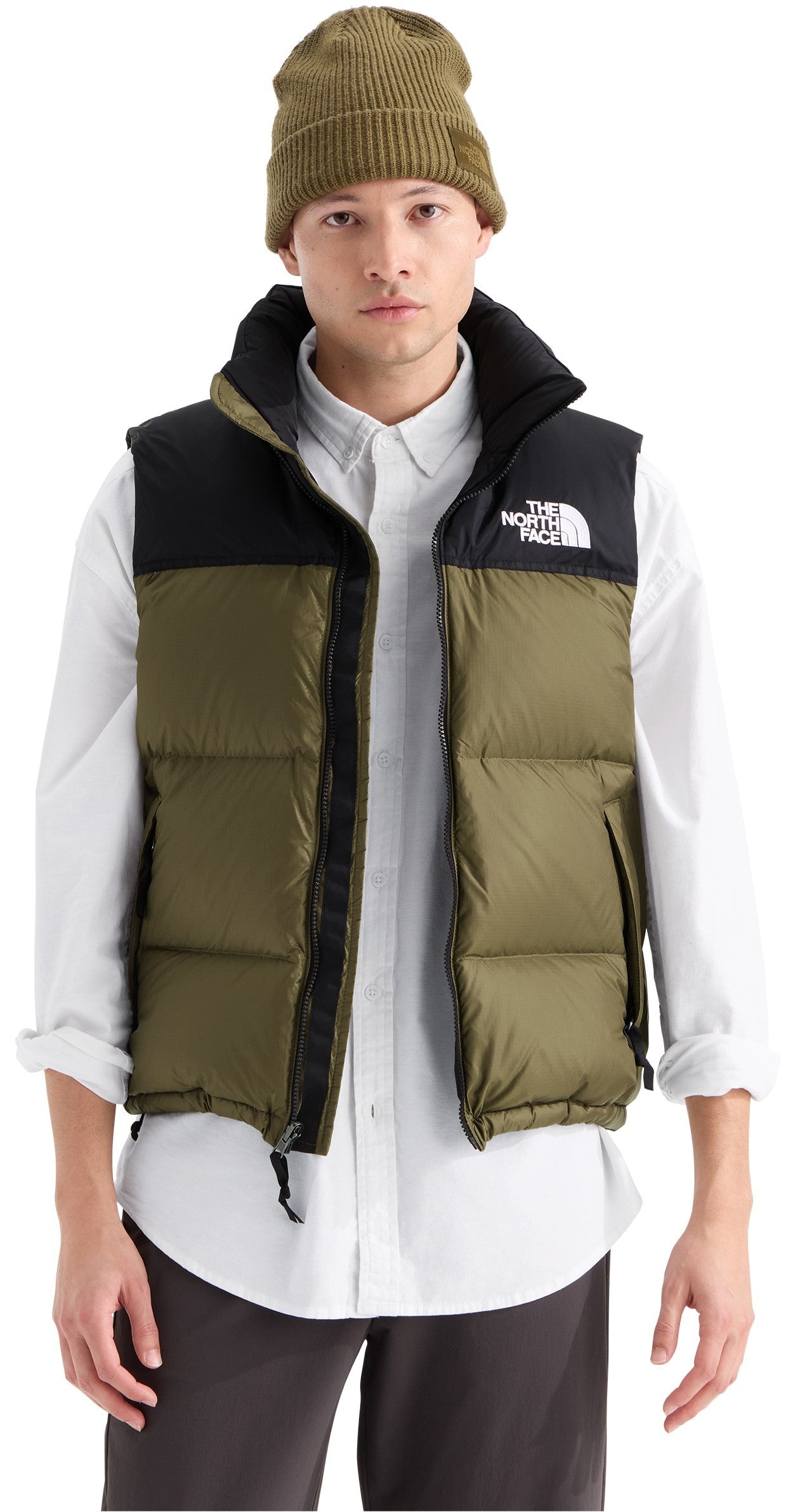 A male model wearing a mustard green beanie, a white button-down shirt with the sleeves rolled up, a two-toned mustard green and black puffy down vest with the brand name printed in white on the upper left chest, and dark grey trousers.