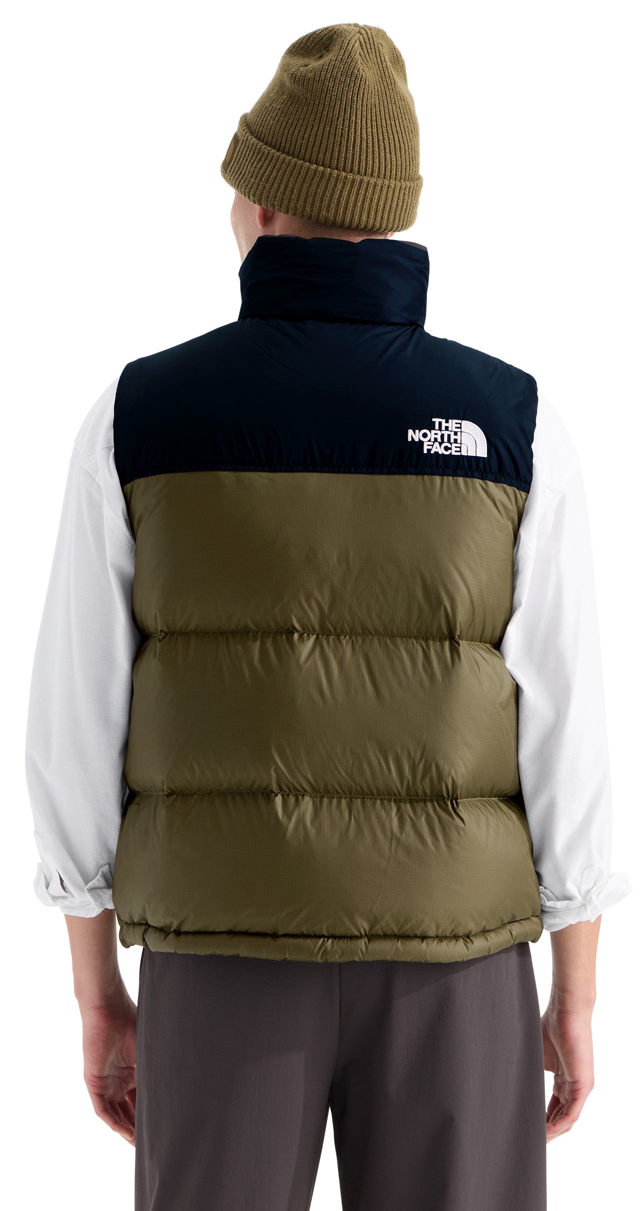A back view of a person wearing an olive green and black puffer vest with white text reading 'THE NORTH FACE' on the upper back, over a white long-sleeved shirt and brown pants, with a matching olive green beanie.