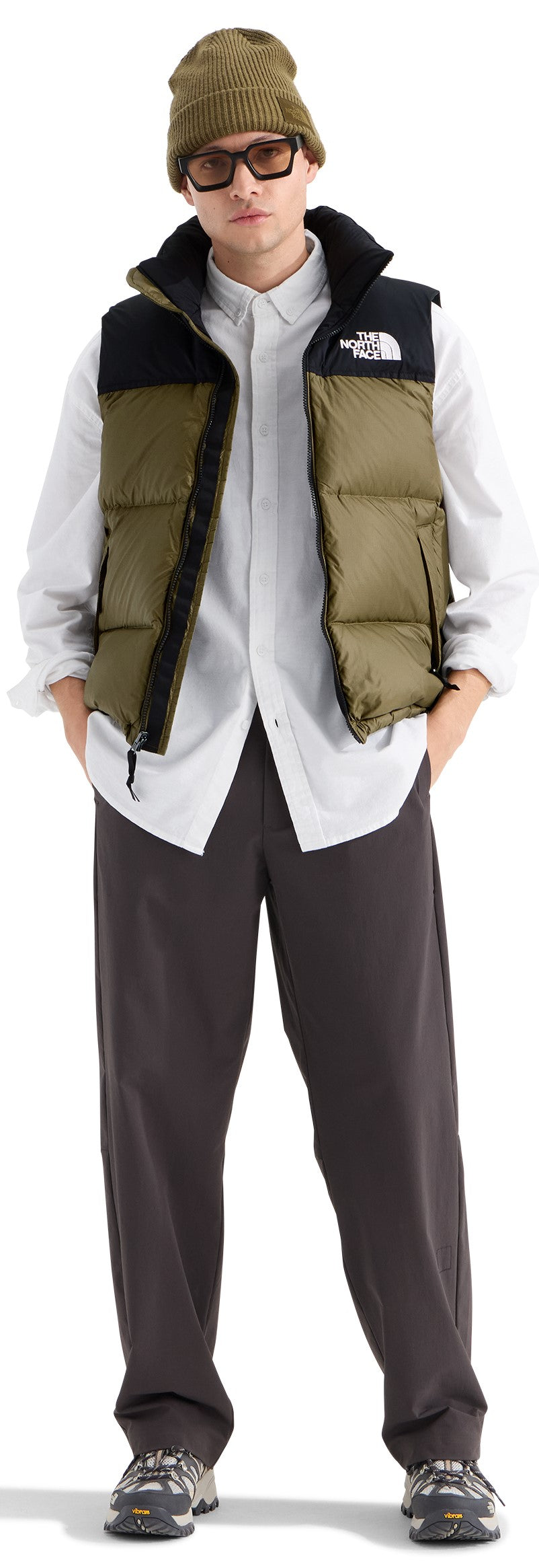 A man wearing a green beanie, dark glasses, a white button-down shirt, a two-toned olive green and black puffer vest, dark grey relaxed-fit trousers, and dark grey sneakers.