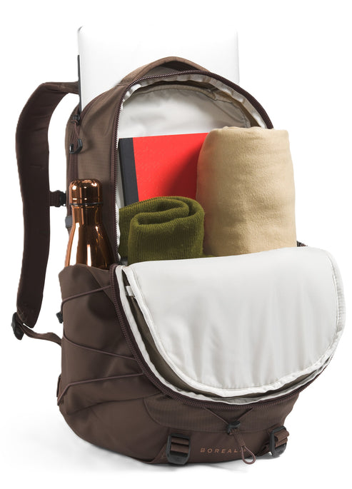 Open dark brown backpack with champagne bottle in side pocket, red notebook, olive green rolled shirt, and rolled light brown blanket inside.