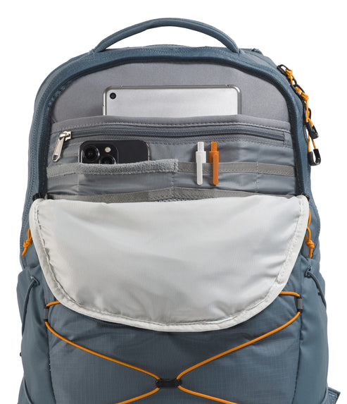 An open, dusty-blue backpack front compartment showing internal storage organization, including a light gray tablet sleeve, a zippered pocket holding a smartphone, two pen loops holding one white and one orange pen, and an external crisscross bungee cord system.