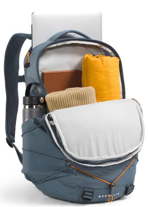 A blue Borealis backpack, shown from a three-quarters front angle with its main compartment fully unzipped and open. The interior is packed with a laptop, a brown notebook, a rolled-up yellow item, a folded beige knit garment, and a water bottle standing in the side pocket. The front of the bag features a zigzagging orange bungee cord system.