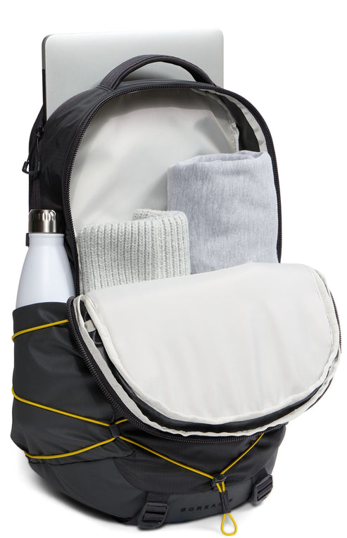 A black backpack with yellow bungee cords on the front, showing an open main compartment filled with folded light gray clothing, a laptop sleeve with a silver laptop inside, and a water bottle in the side pocket.