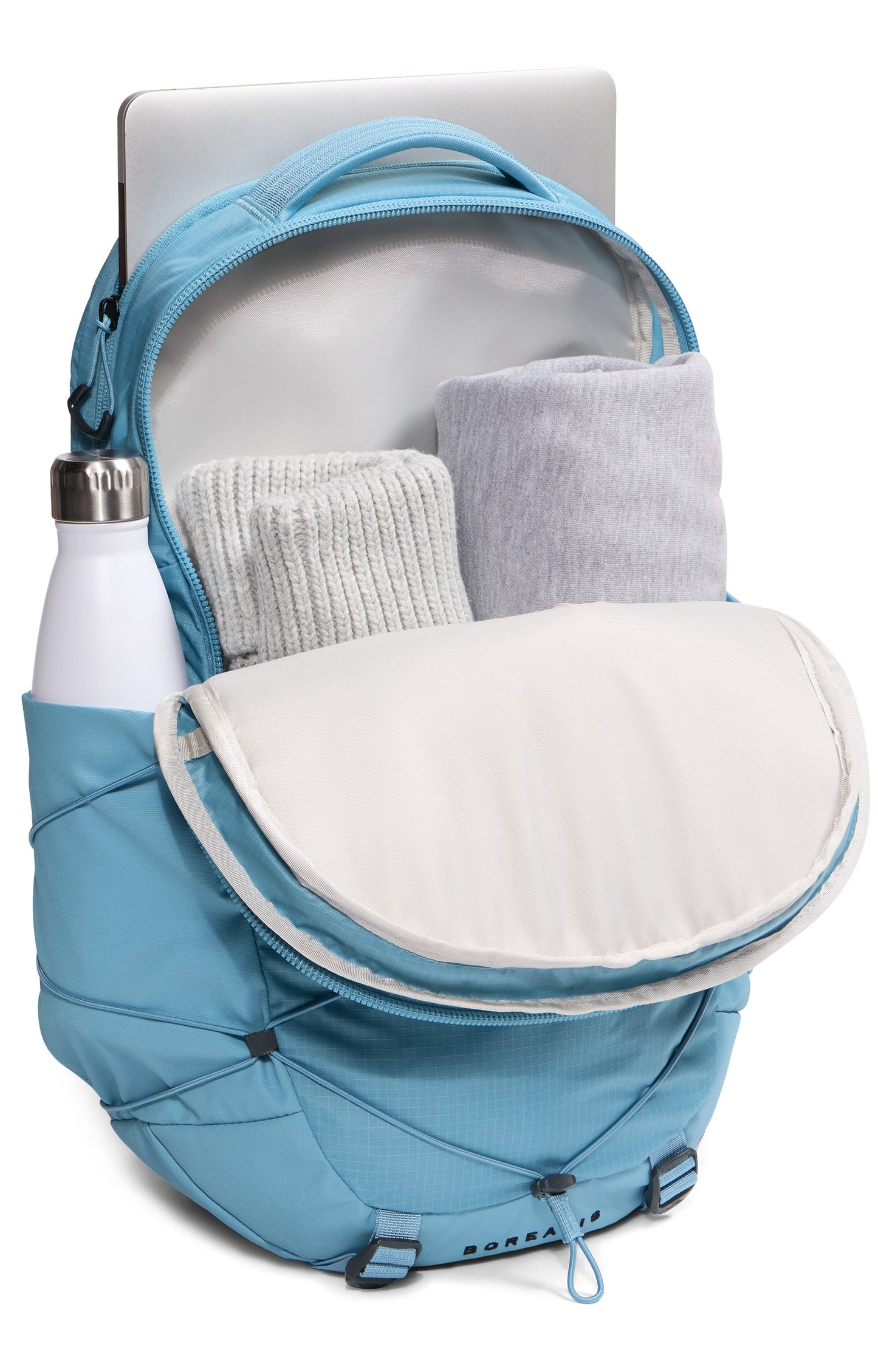 A light blue backpack with its main compartment partially open, containing rolled-up grey clothing items and a laptop in the rear sleeve, with a white water bottle stored in the side pocket.