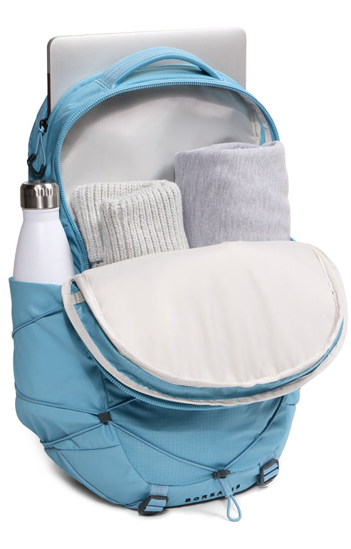 A light blue backpack with its main compartment partially open, containing rolled-up grey clothing items and a laptop in the rear sleeve, with a white water bottle stored in the side pocket.