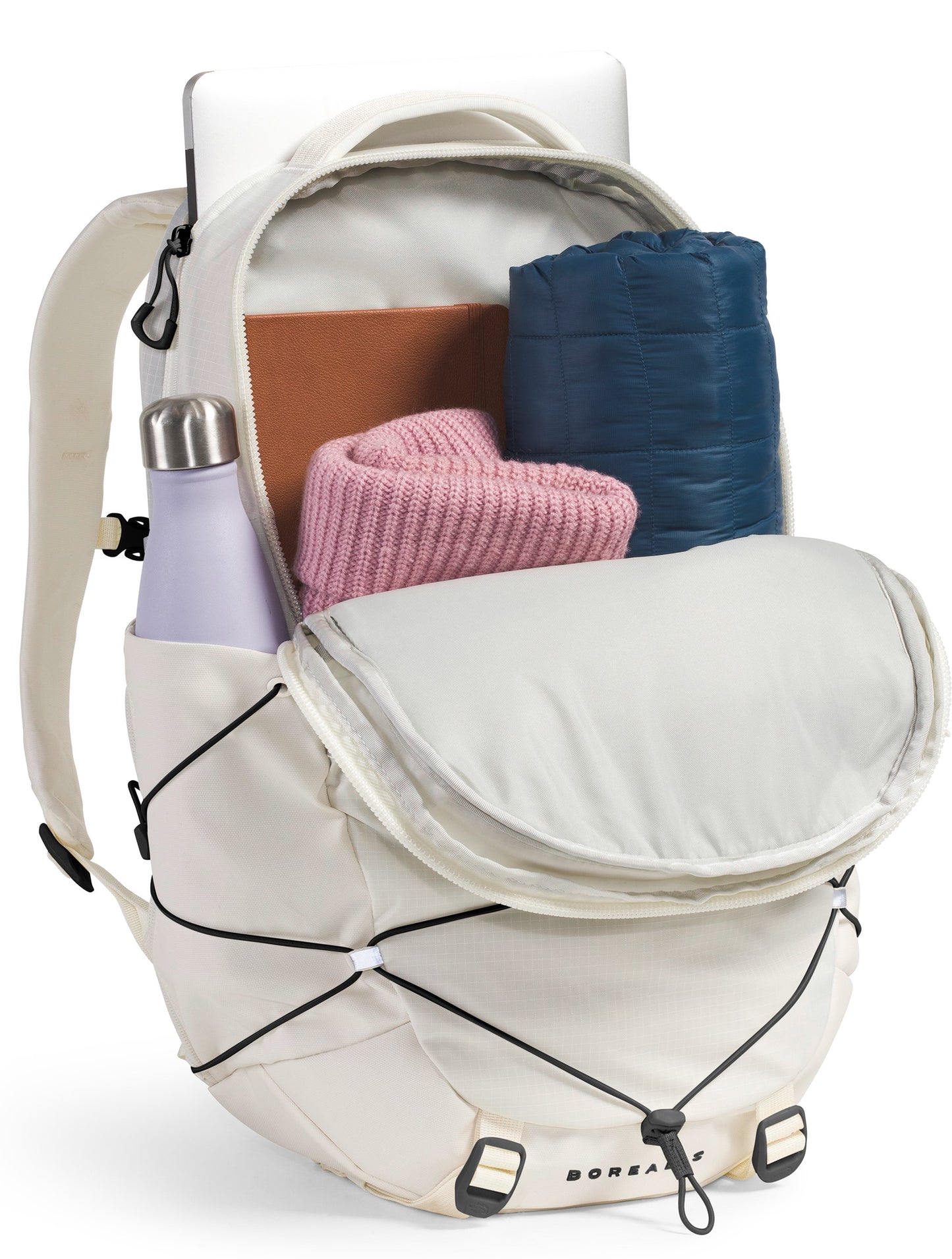A light beige, open backpack with a laptop, a light purple water bottle, a folded pink sweater, and a folded dark blue jacket inside, featuring an external black bungee cord system on the front.