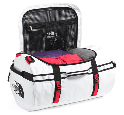 Open white duffel bag with black and red straps and "THE NORTH FACE" printed on the side, partially filled with clothing and showing a zippered mesh compartment on the inside of the top flap.