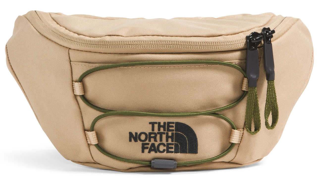 A tan-colored waist pack with a horizontal green bungee cord system, a black zipper closure with dual matching pulls, and the text THE NORTH FACE printed in black.