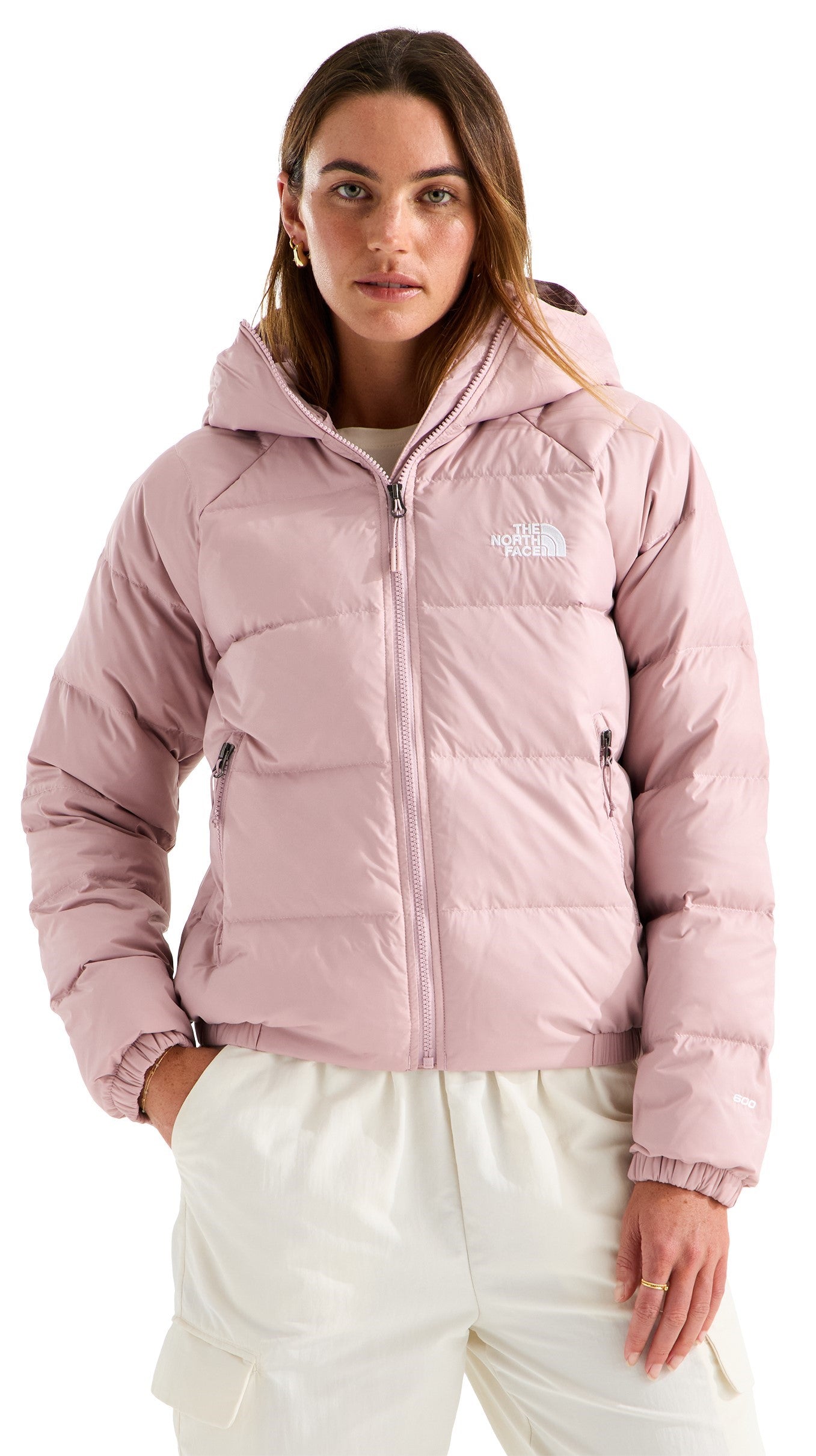 A woman wears a light pink, hooded, quilted puffer jacket with a full-length front zipper and the text "THE NORTH FACE" embroidered on the upper left chest, paired with cream-colored cargo-style pants.