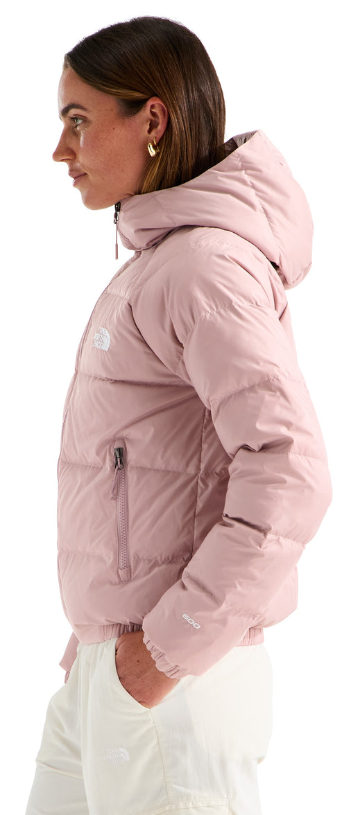 A woman seen from the side profile, wearing a pale pink, hooded, quilted, long-sleeved down jacket with a front zipper and light-colored trousers.