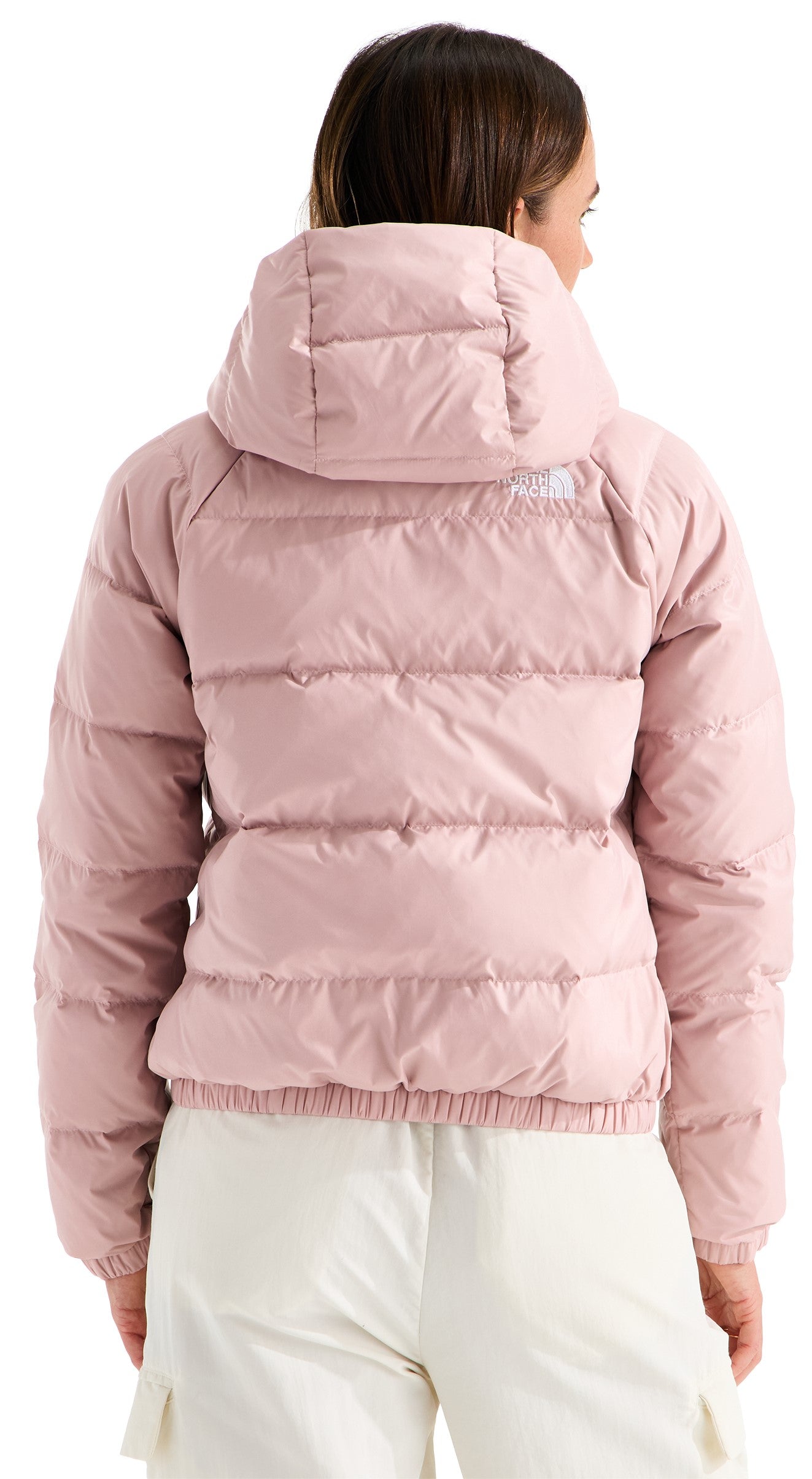 A back view of a woman wearing a light pink, hooded, quilted, insulated down jacket over light-colored pants.