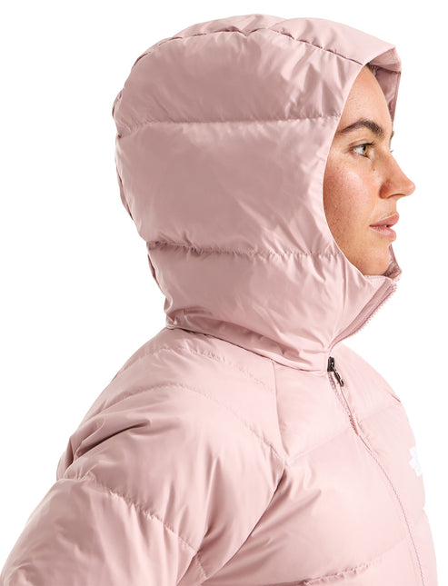 A close-up side profile view of a woman wearing a light pink, hooded, quilted, insulated winter jacket, with the hood pulled up over her head.