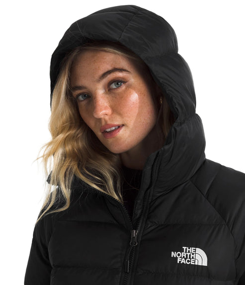A fair-skinned woman with blonde hair wearing a black puffer jacket with an insulated hood pulled up, the text THE NORTH FACE is embroidered on the chest.