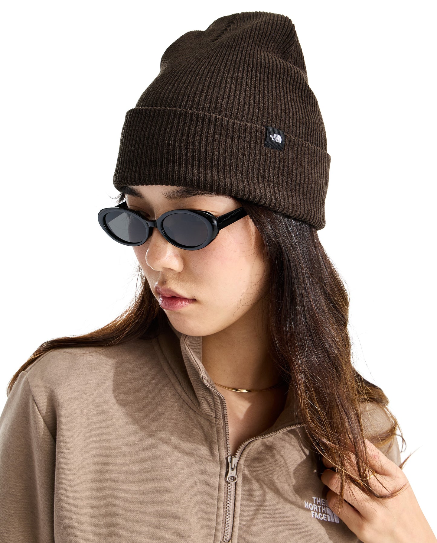 A young woman wearing a dark brown ribbed beanie with a small rectangular logo tag on the cuff, black oval sunglasses, and a tan quarter-zip fleece jacket.