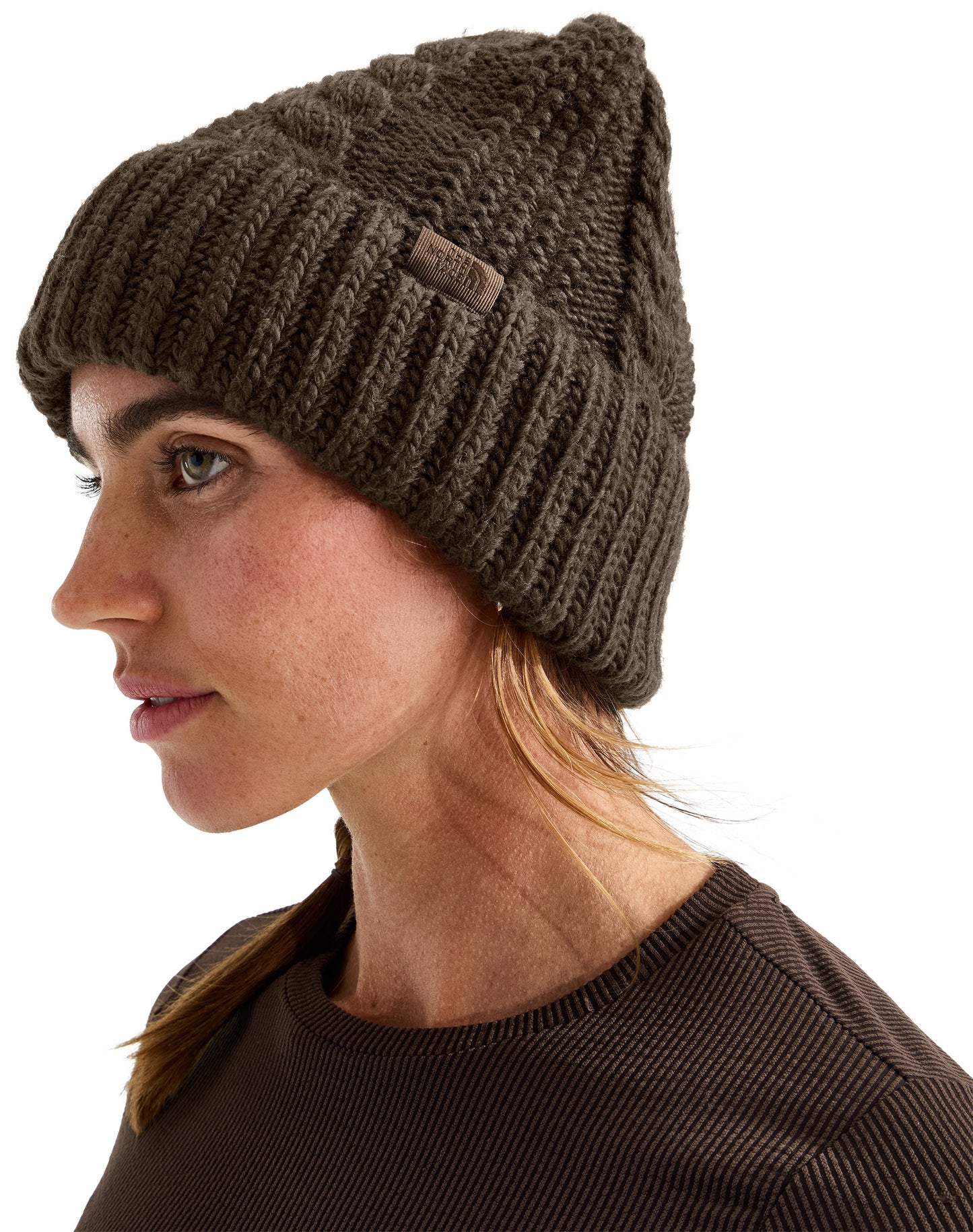 A woman in profile view wearing a dark brown chunky knit cuffed beanie with a small rectangular fabric label attached to the cuff.