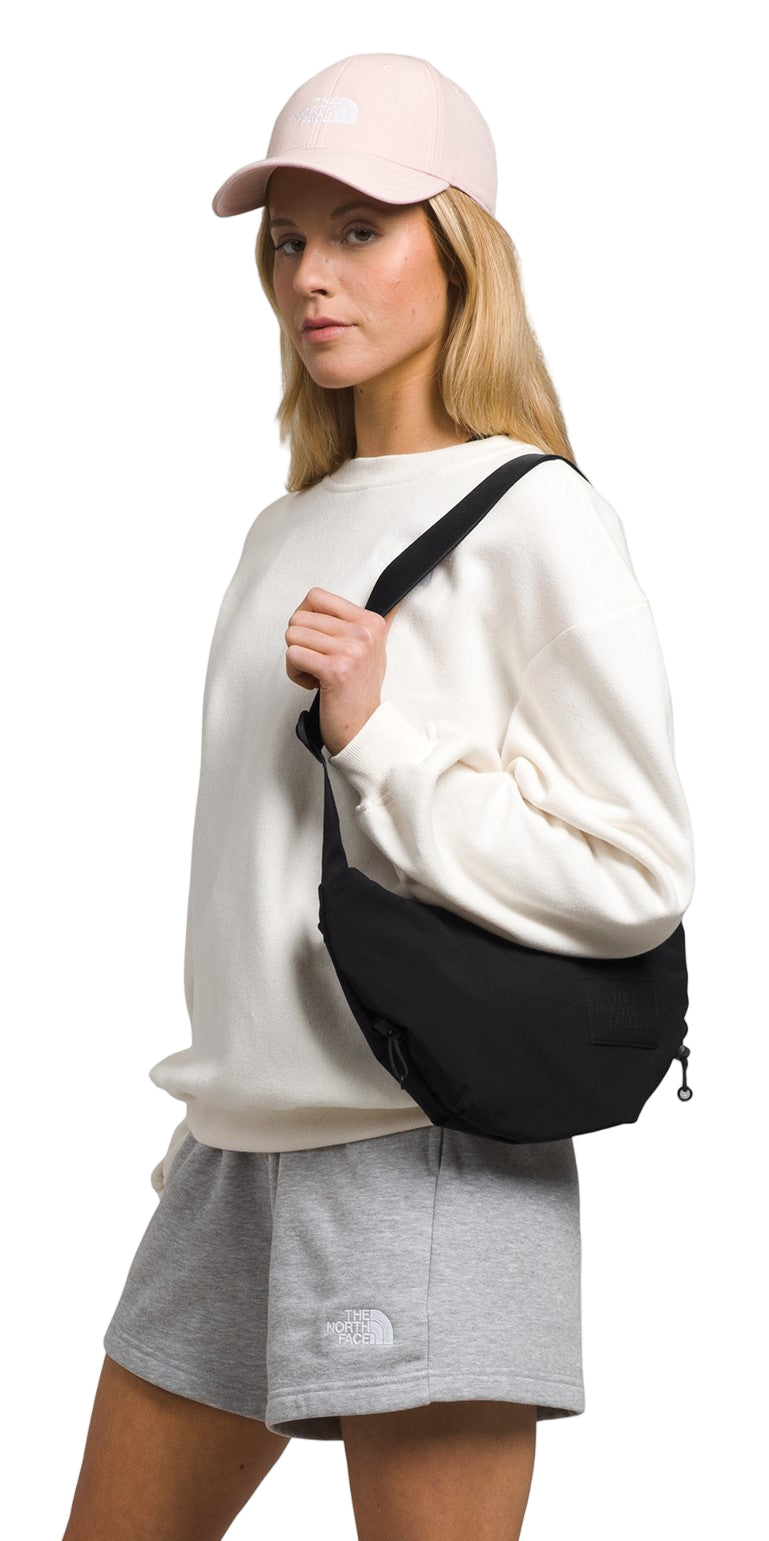 A woman wearing a pink hat, white sweatshirt, and gray shorts has a black shoulder bag on.