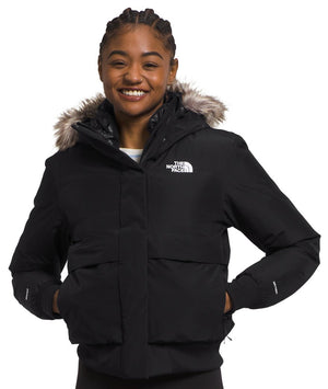 A smiling person wearing a black, waist-length insulated winter bomber jacket with a hood featuring a faux-fur trim and a small white brand logo printed on the upper left chest.
