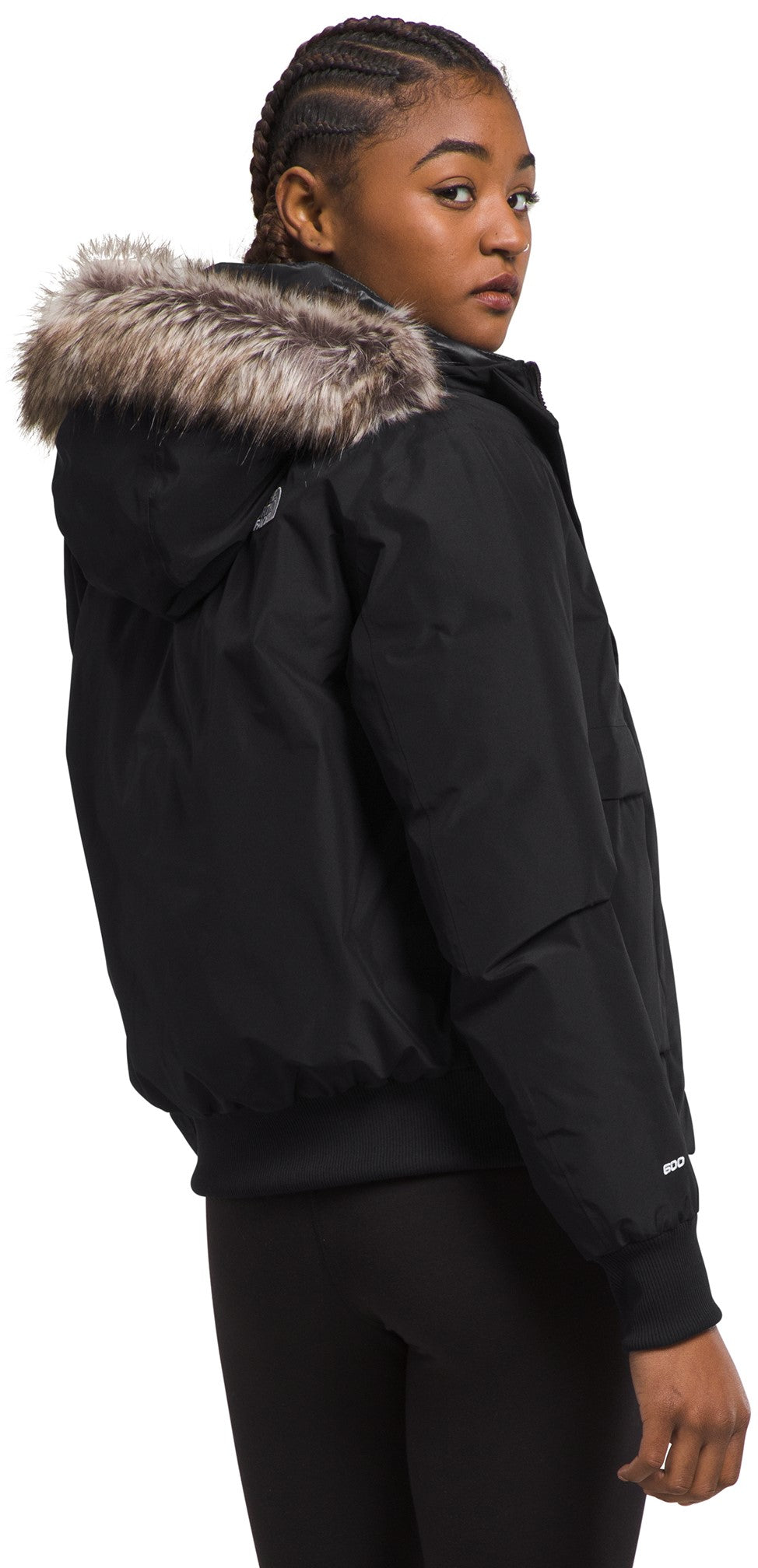A young woman wearing a black, hooded, down-filled winter bomber jacket with a fur-trimmed hood, seen from a side-rear angle, looking back at the camera.