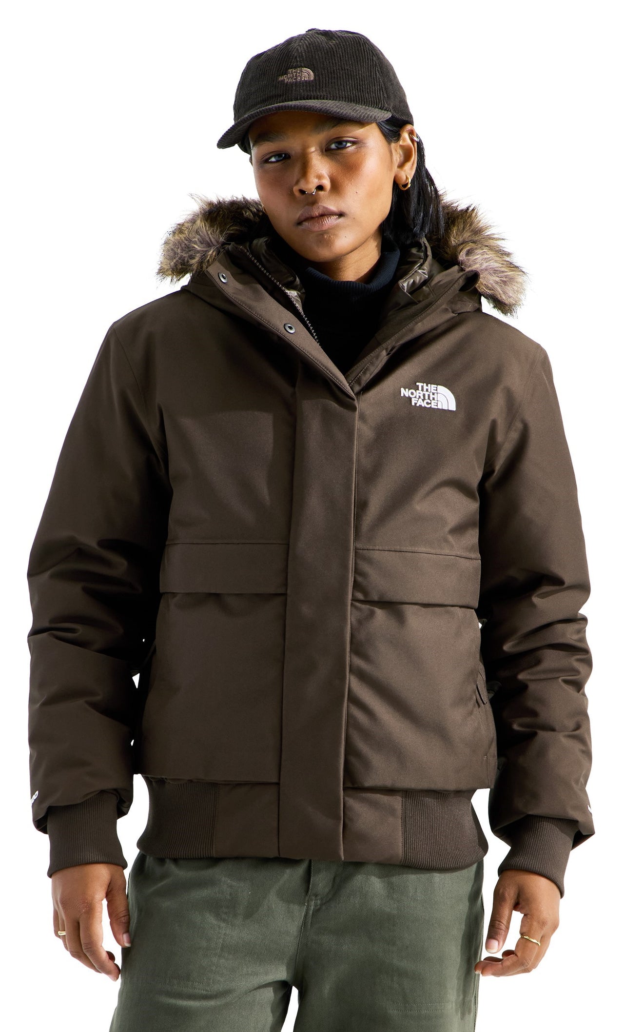 A person wearing a dark brown, hooded bomber-style jacket with a faux-fur trim on the hood, ribbed cuffs and hem, and a white logo on the chest, paired with a dark brown baseball cap.