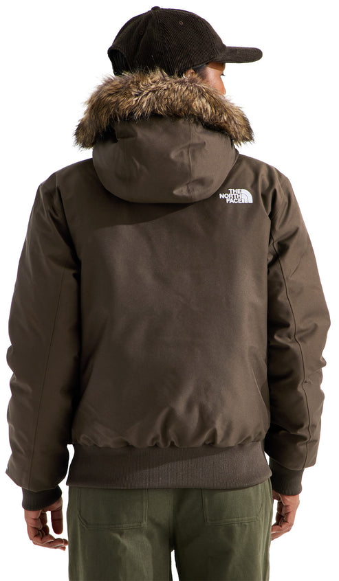 A person viewed from the back wearing a dark olive green, waist-length, hooded winter jacket with a brown faux fur trim around the hood, dark corduroy baseball cap, and light olive green pants, with the brand name logo printed on the back right shoulder.