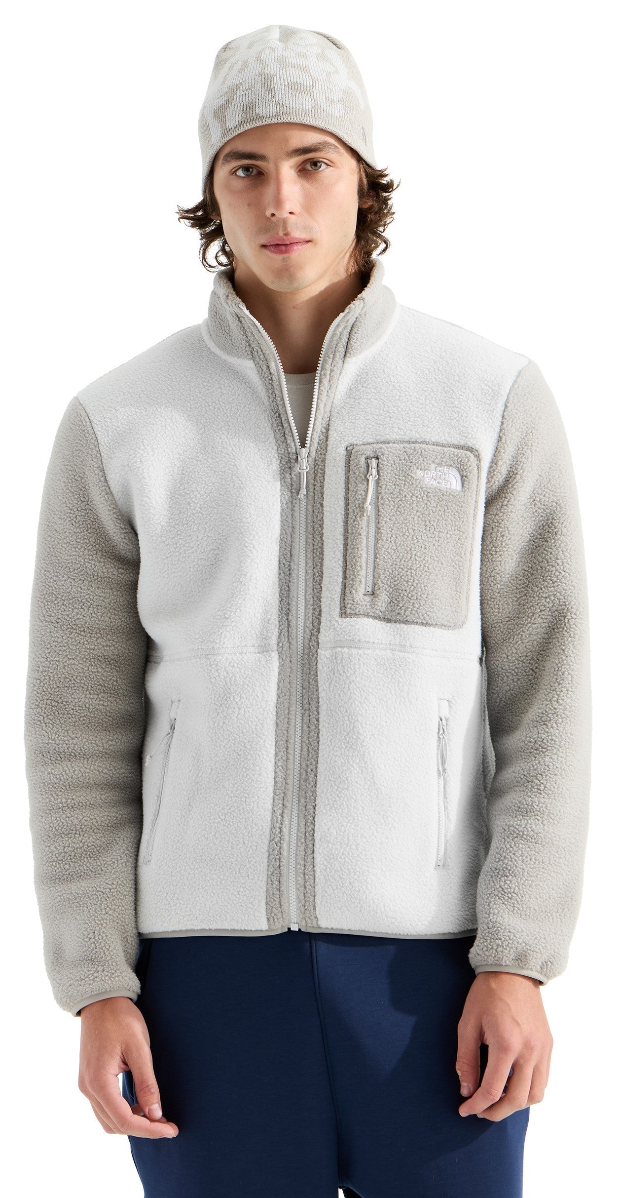 A man wearing a white, high-collared, zip-up fleece jacket with textured gray sleeves, a gray chest pocket, and a matching gray patterned beanie.
