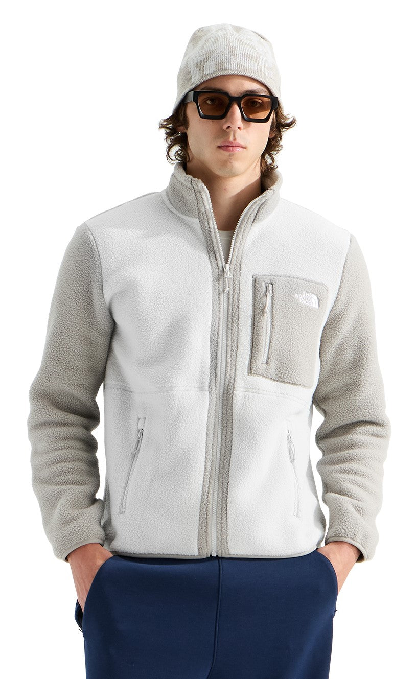 A person wearing a light grey zip-up fleece jacket with a textured sleeve design, a chest pocket with a logo, a matching beanie, and dark-rimmed sunglasses, styled over dark blue pants.
