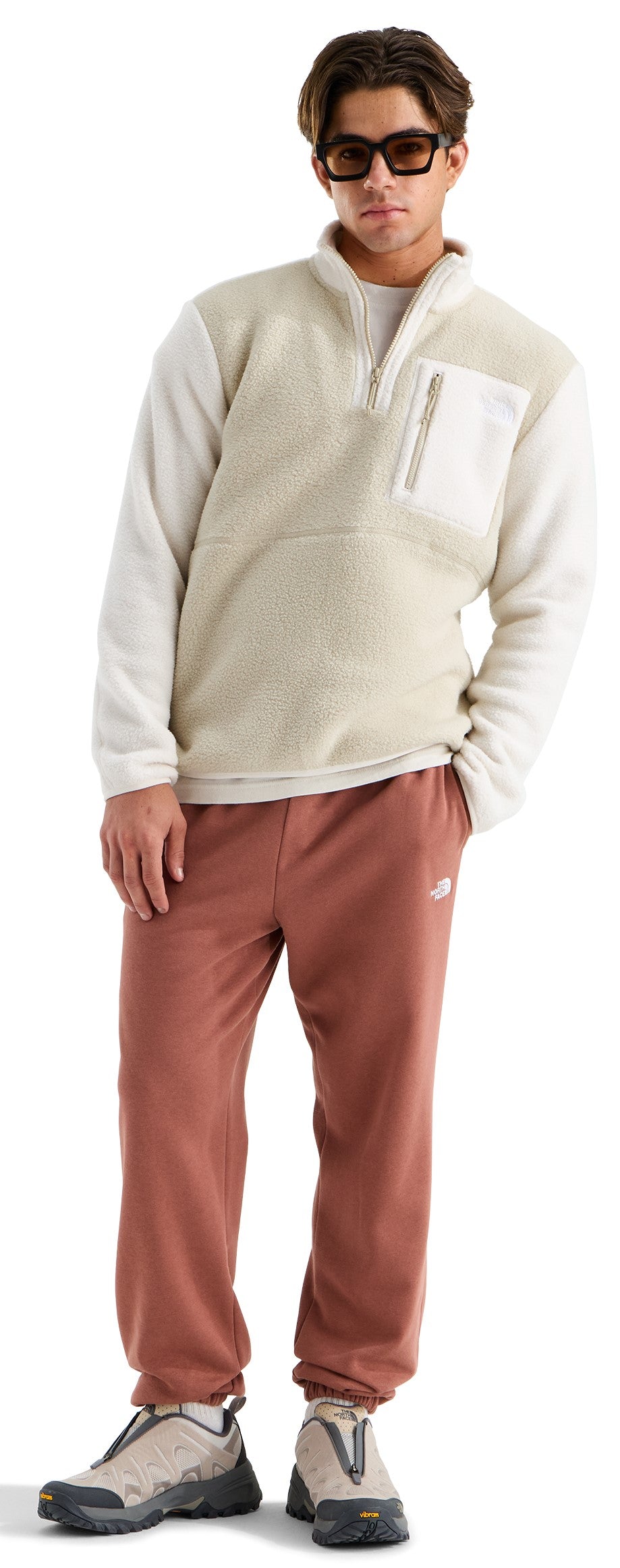 A man wearing a cream-colored, fuzzy, quarter-zip fleece pullover with a vertical zippered chest pocket, rust-colored sweatpants, and tan, trail-style sneakers with black trim. He has dark hair and is wearing dark-framed sunglasses.