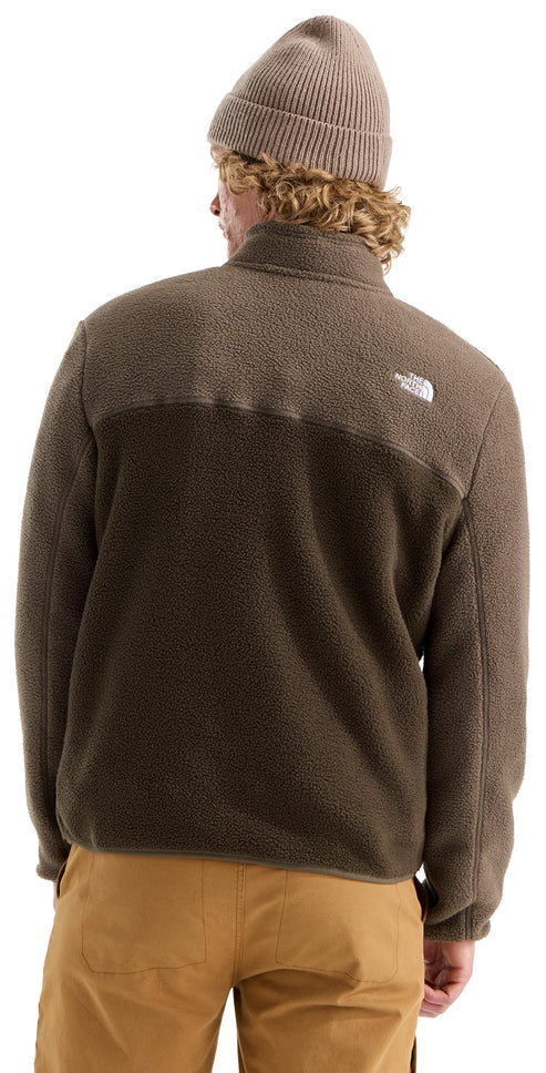 A back view of a person with blonde, curly hair wearing a tan knit beanie and a brown, textured fleece jacket with a high collar and a small logo on the upper right shoulder, paired with tan pants.