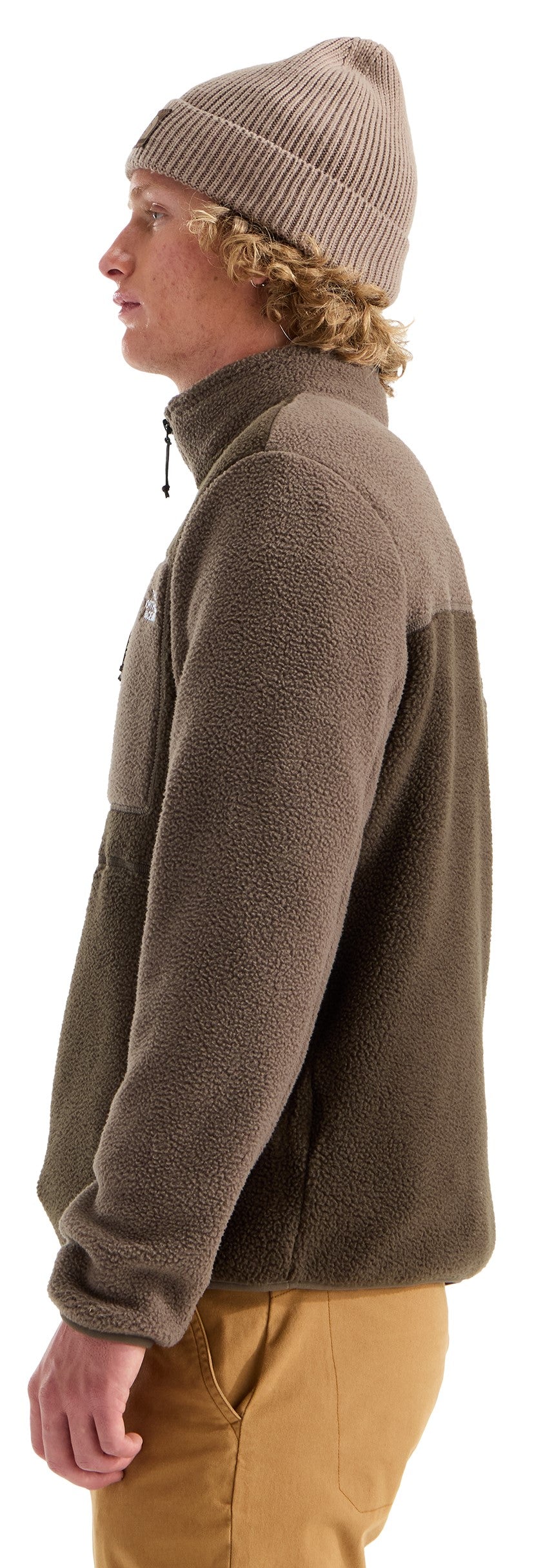 A side view of a person with blonde curly hair wearing a tan ribbed beanie, a taupe-colored quarter-zip fleece jacket with a small chest pocket, and tan pants.