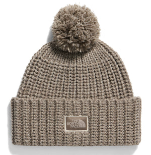 A beige knit beanie with a ribbed texture, a large pom-pom on top, and a rectangular patch in the center with The North Face logo.